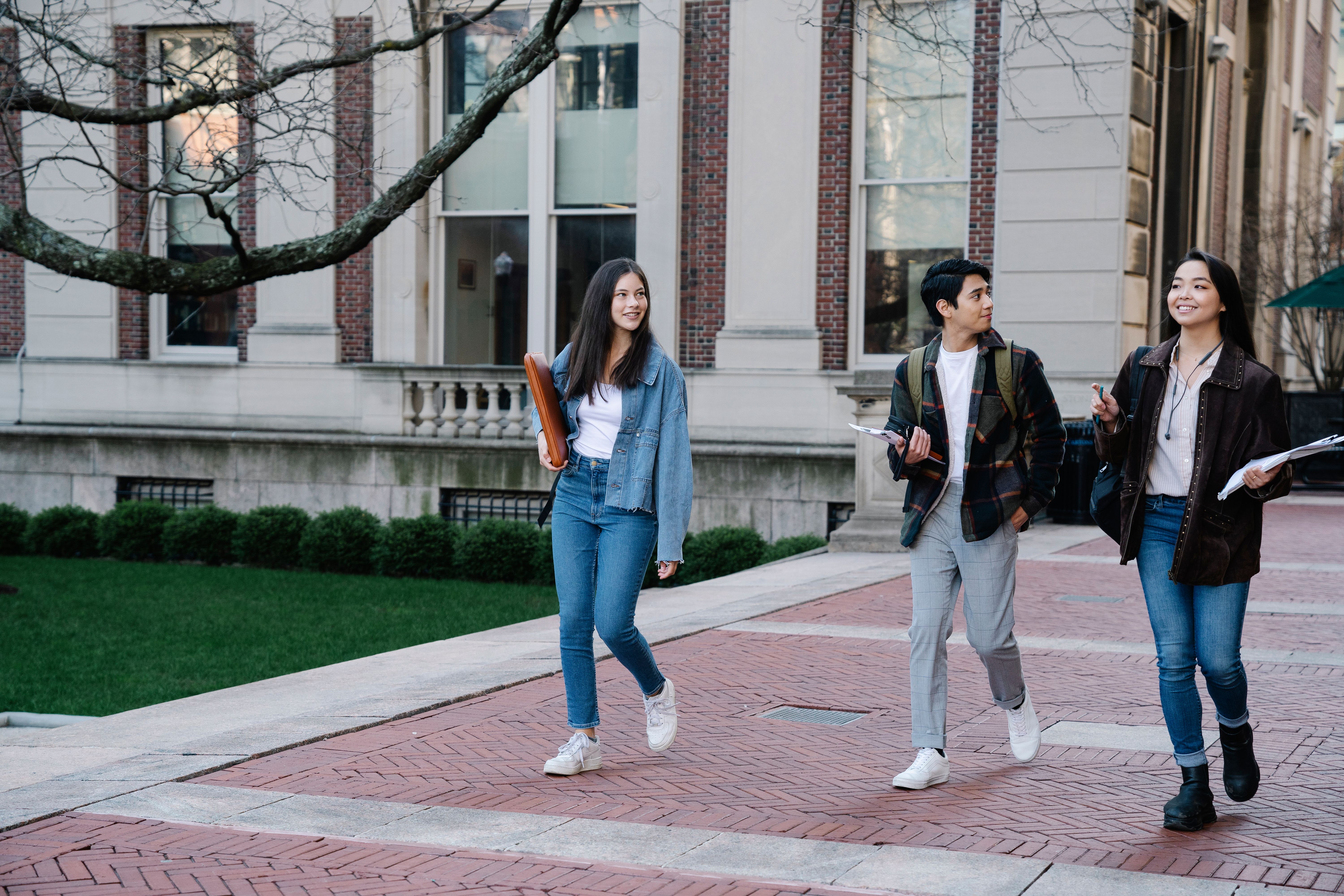 Three university students walk and talk along a campus path carrying books and study materials