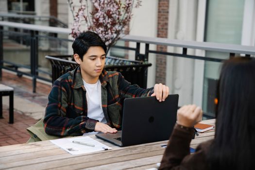 Young man using a laptop on a wooden table, working remotely outdoors. Urban setting.