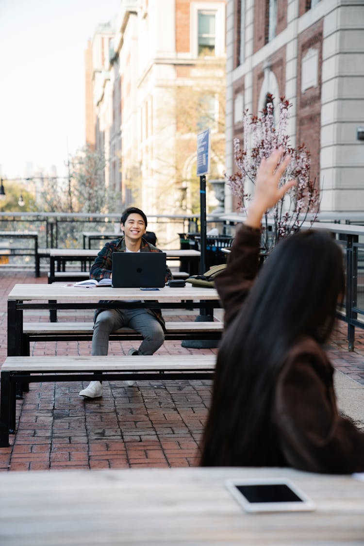 Woman Waving To A Man Sitting Behind Her With A Laptop 