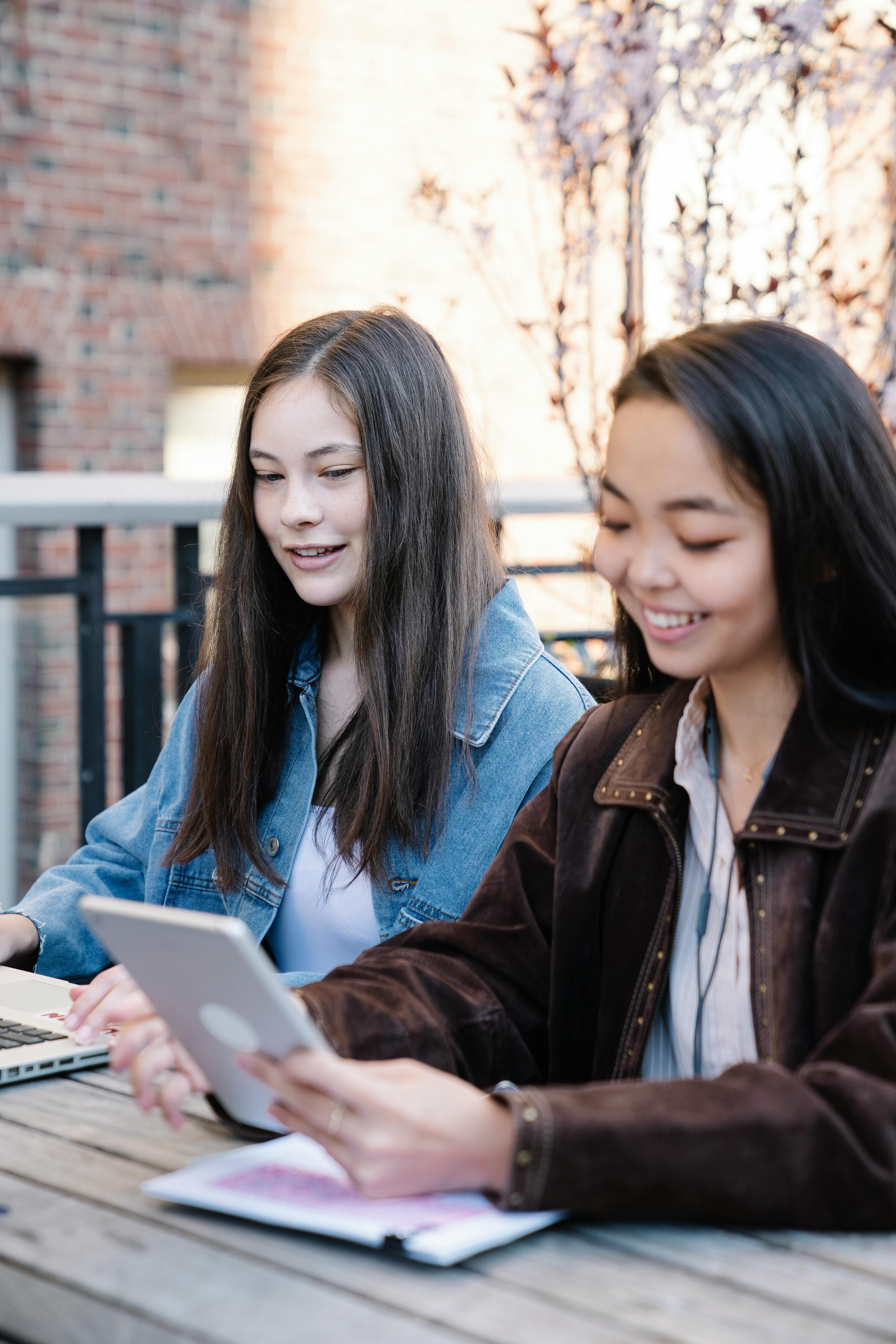 Two teenage girls studying and smiling at a table outdoors, using a tablet and laptop.