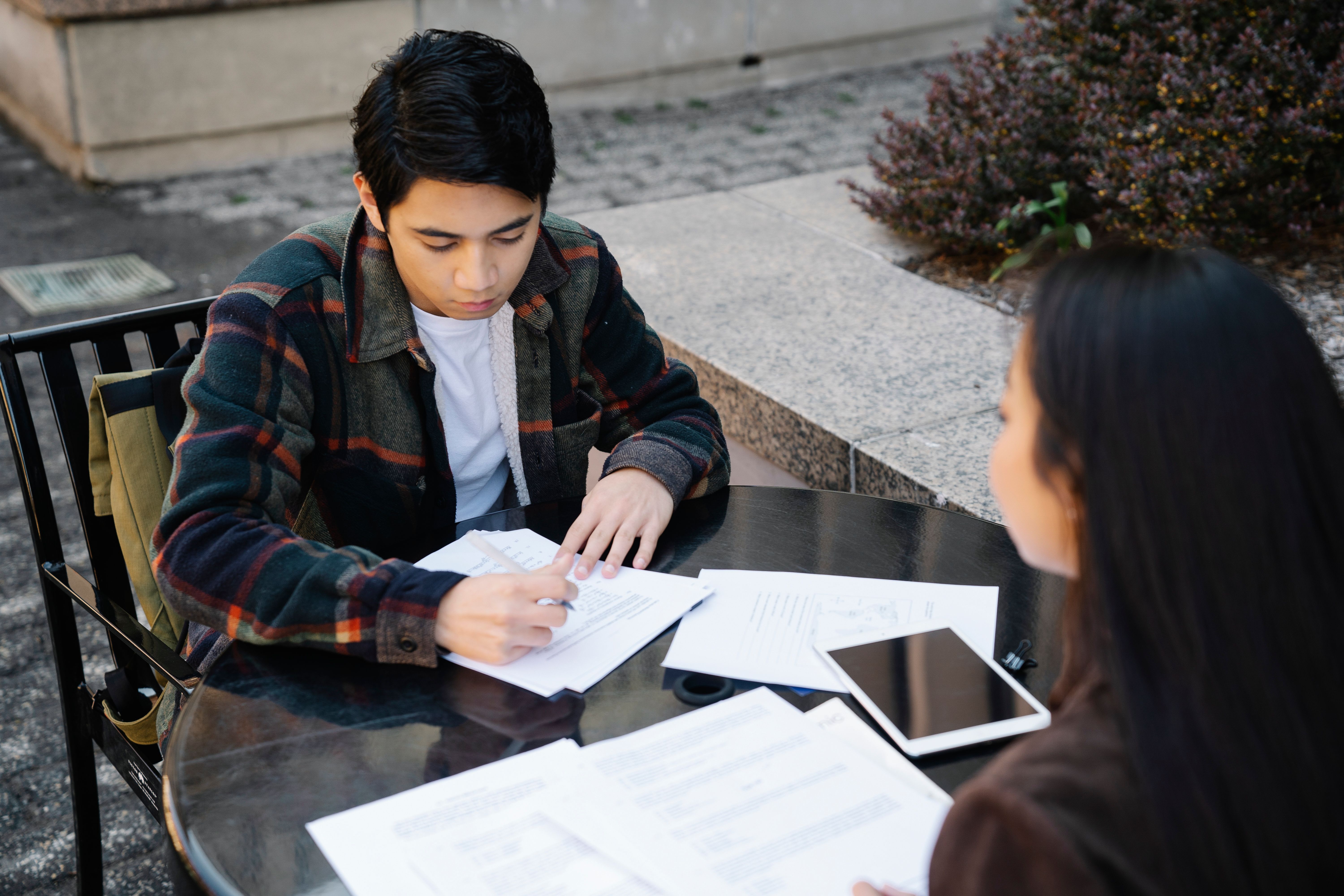 Two students are working together at a table outside, discussing and writing on papers.