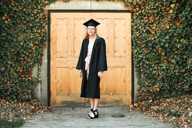 Positive University Graduate In Academic Cap Standing Near Wooden Doors