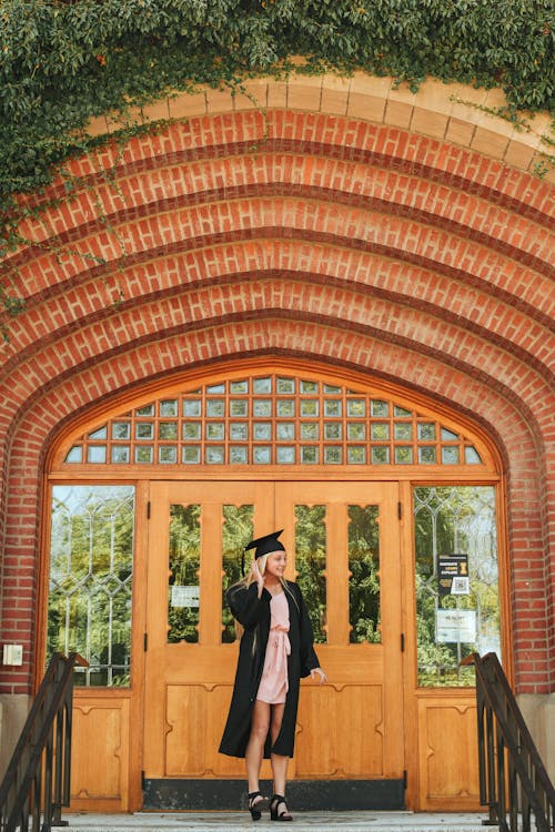 Full body of young female student wearing graduation cap and black cape standing near door of old