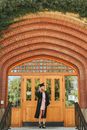 Full body of young female student wearing graduation cap and black cape standing near door of old