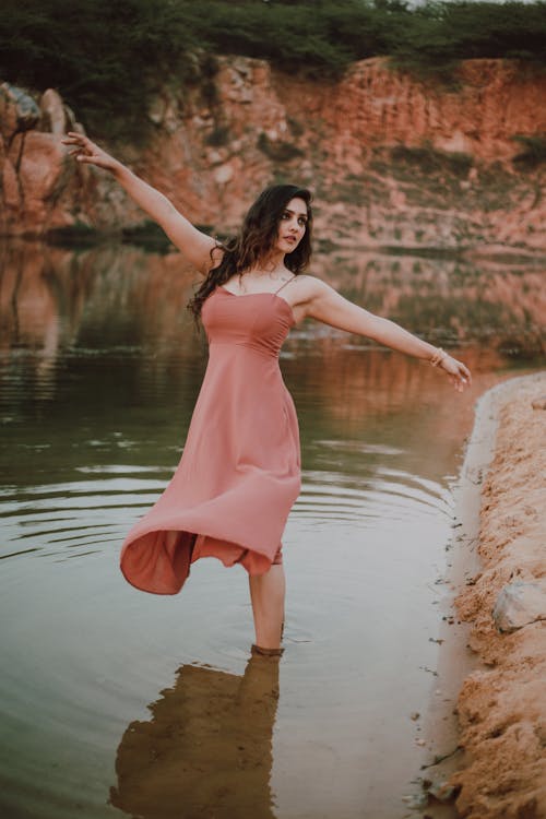 Free Slim graceful female with wavy dark hair wearing summer dress dancing in tranquil water of river near sandy coast Stock Photo