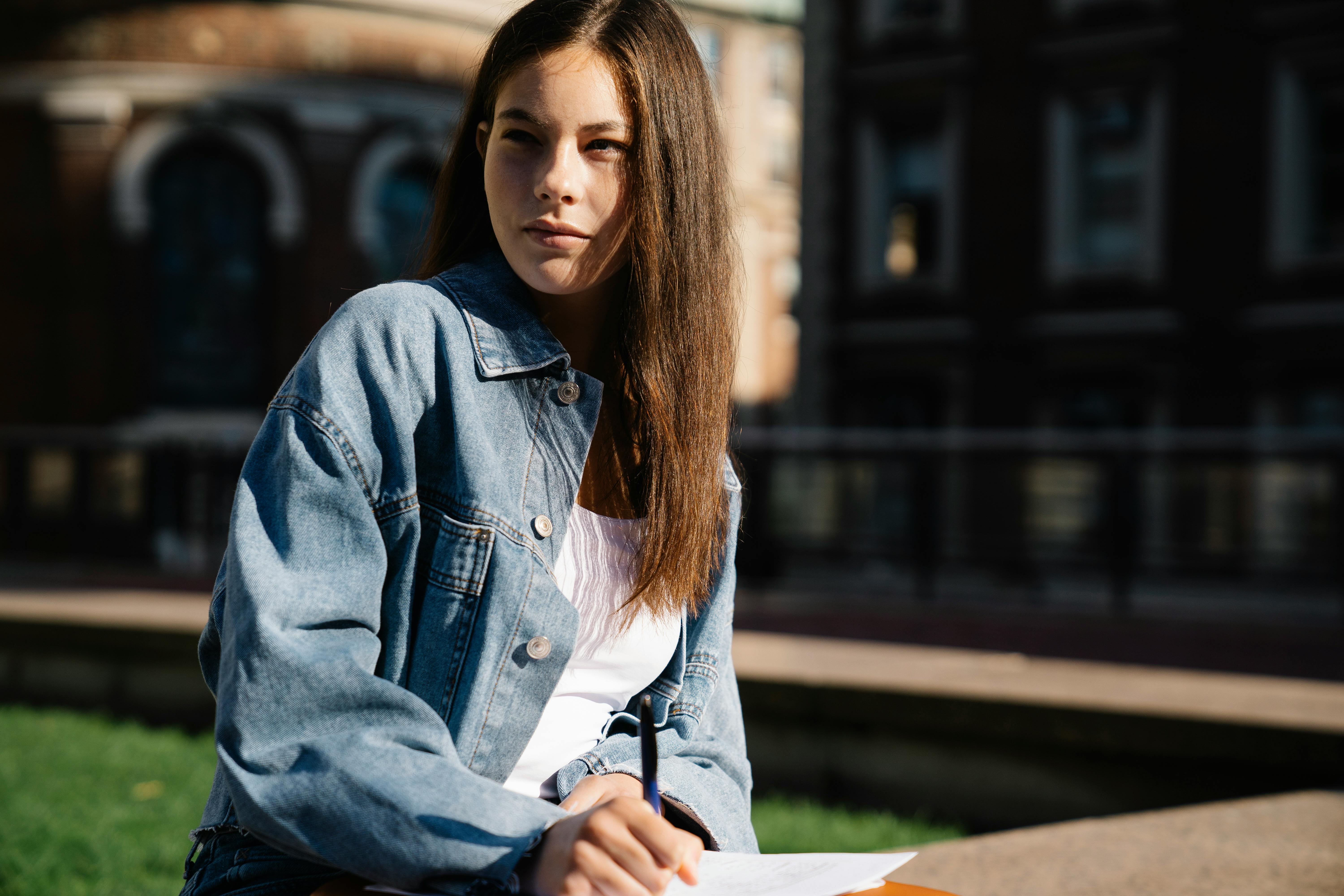 A young woman in a denim jacket studying outdoors in the sunlight, with focus and intent.