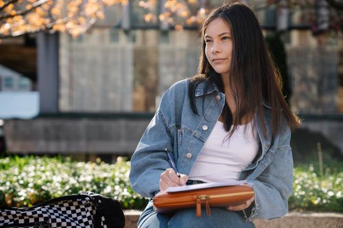 Free A young woman in a denim jacket writing outdoors in a university campus setting. Stock Photo