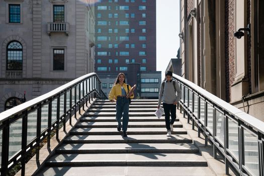 Two young adults walking down university steps in an urban setting, holding notebooks.