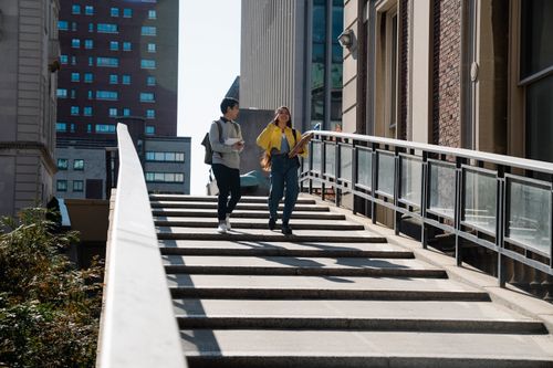 Free Two students walk down stairs on a sunny urban college campus, engaged in conversation. Stock Photo