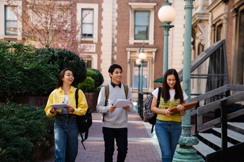 Free Three young adults walking and talking on a sunny campus day, holding notebooks and backpacks. Stock Photo