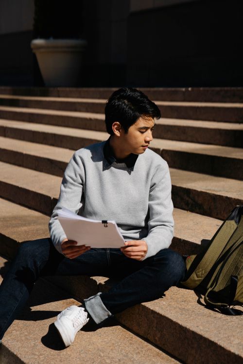 Free A young man sits on steps outdoors, holding papers and a backpack, in casual attire. Stock Photo