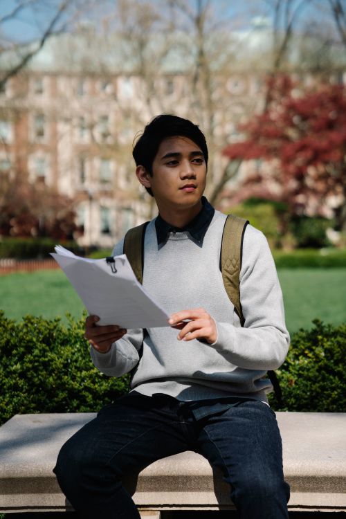 Free A focused young adult student takes notes on a bench in a serene campus environment. Stock Photo