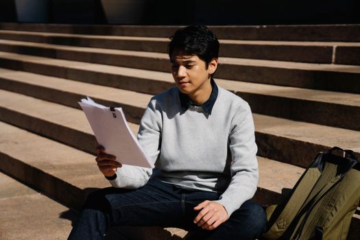 A young man sitting on stairs outdoors, studying papers in the sunlight.