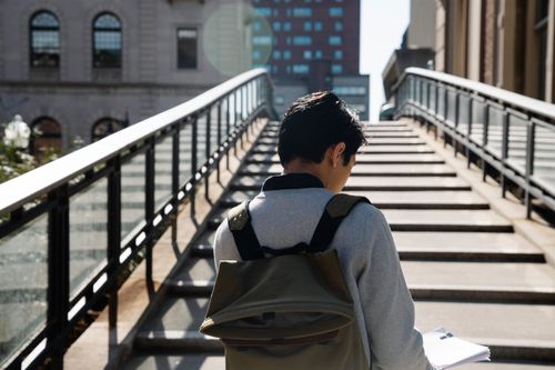 Free A young man with a backpack reads papers on a city stairway under bright sunlight. Stock Photo
