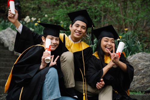 Free Three college graduates in caps and gowns celebrating success outdoors with diplomas. Stock Photo