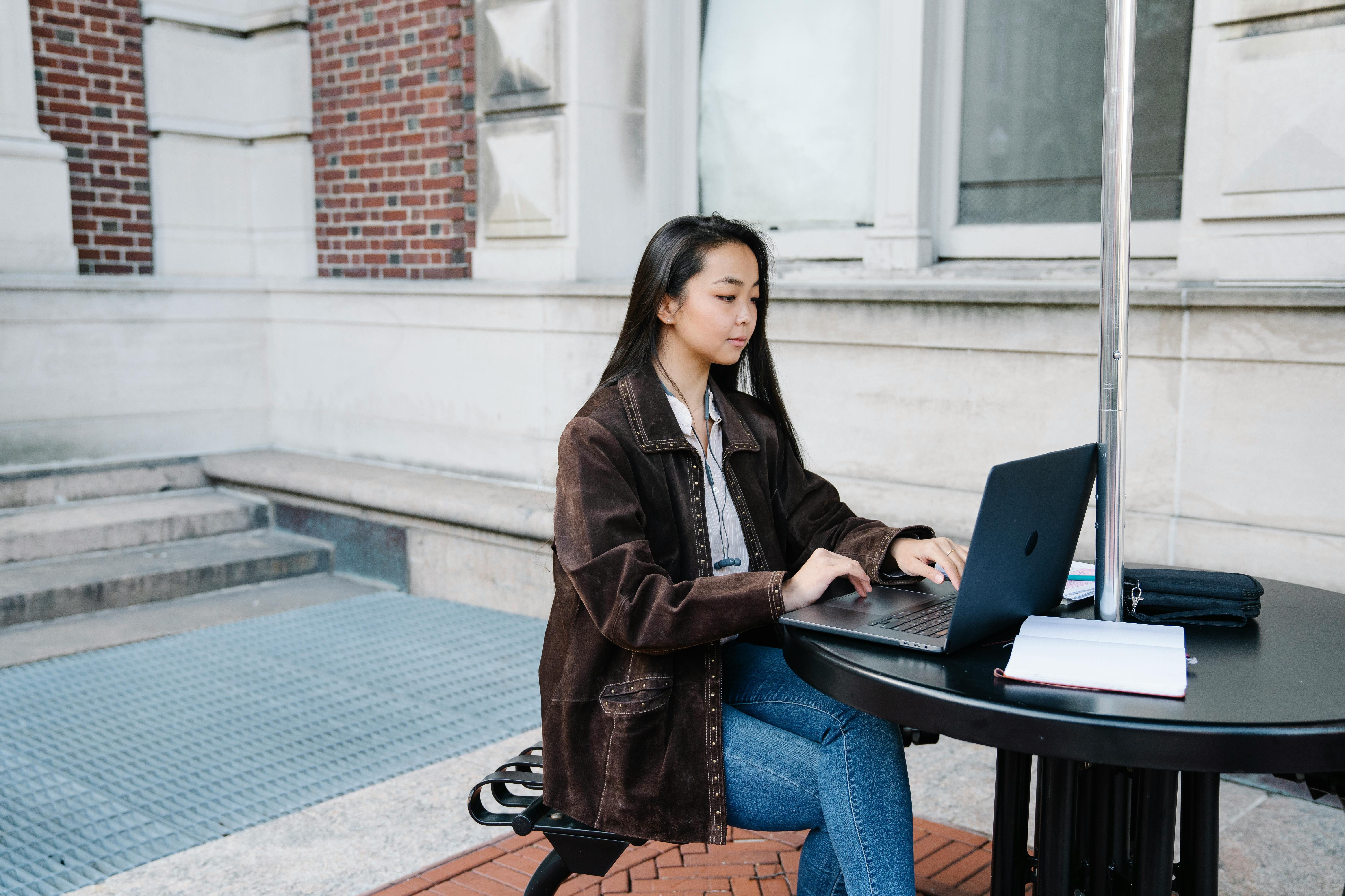 Young woman sits at an outdoor table using a laptop, focused on work.