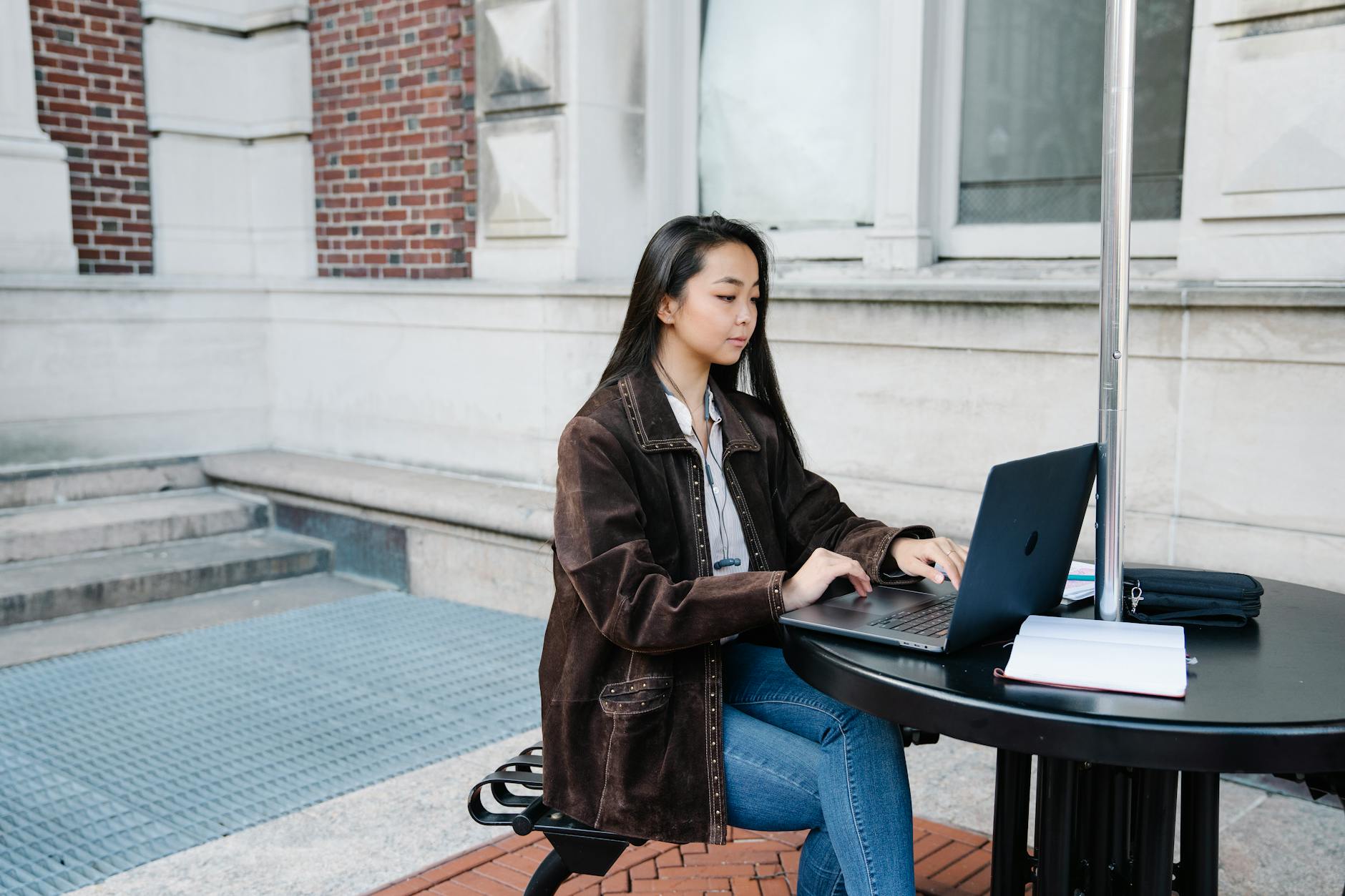 Young woman sits at an outdoor table using a laptop, focused on work.