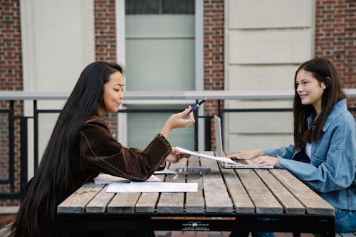 Young Women Sitting at a Table Outdoors Looking at Notes and Using a Laptop 