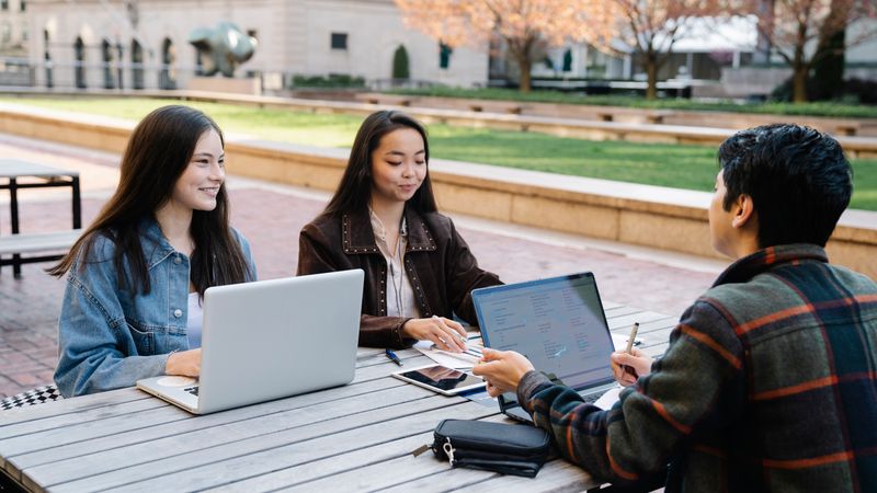 A group of students studying and using laptops together outside in a college campus setting.