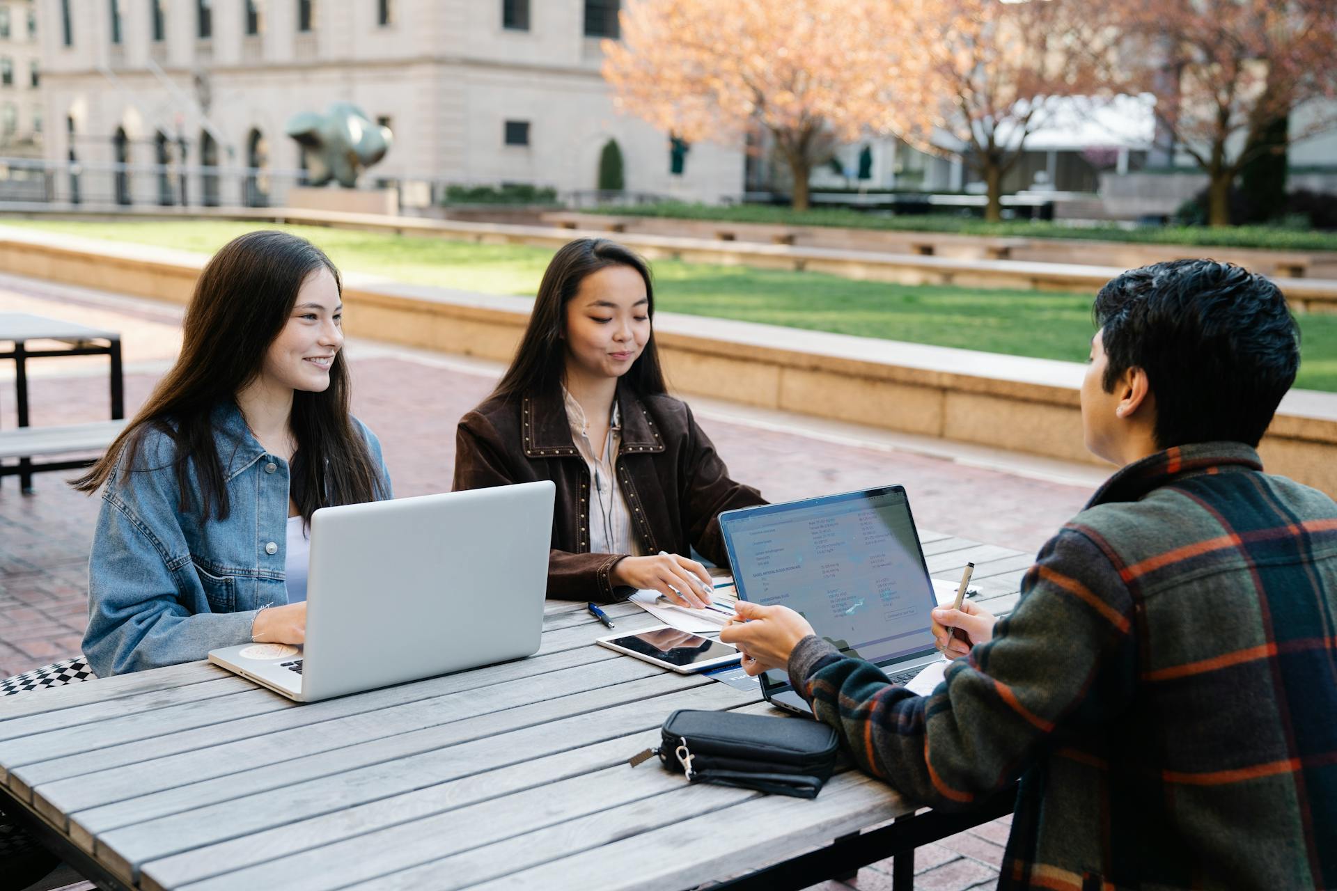 A group of students studying and using laptops together outside in a college campus setting.