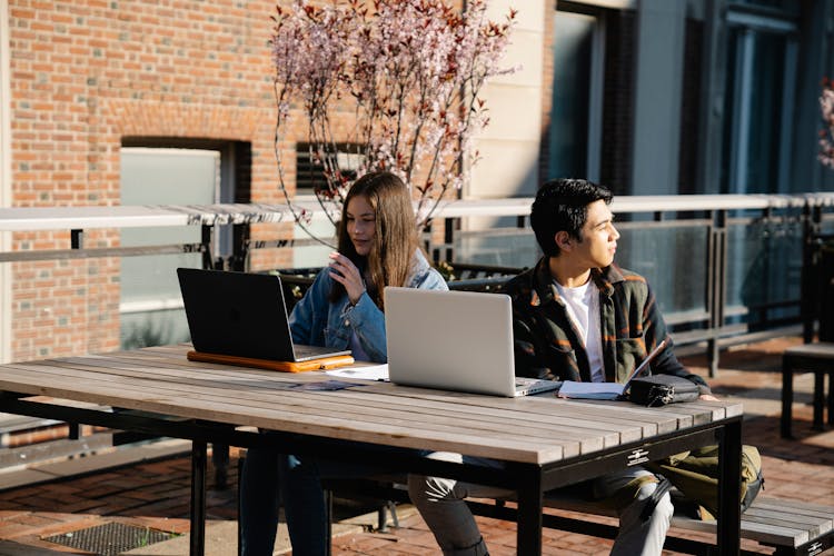 Students Sitting At Table Outdoors Working On Laptops