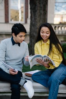 Two college students studying with books on a campus bench, engaged in learning and conversation.
