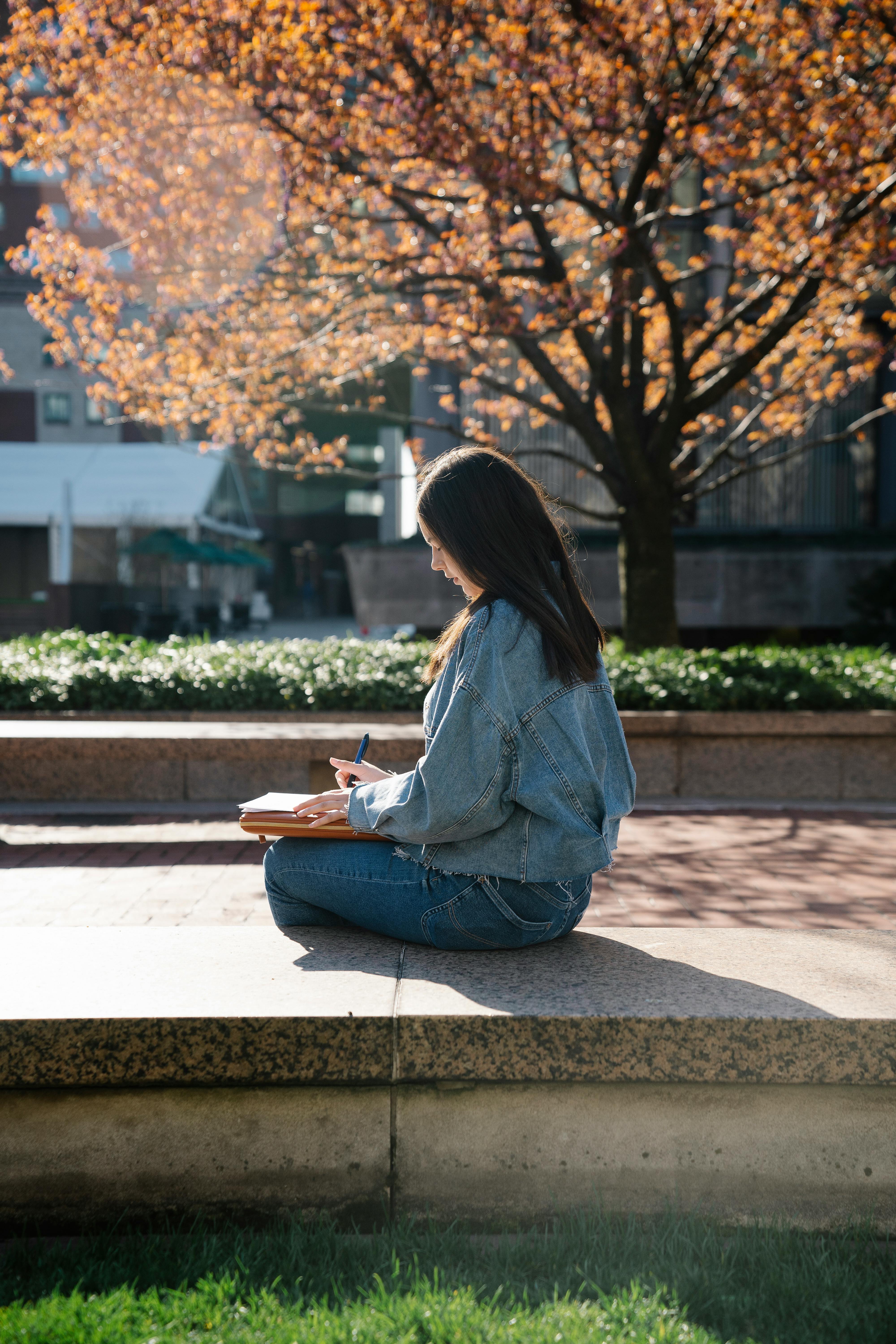 A young woman in casual attire writes in a notebook while sitting on a ledge in a park during a sunny day.
