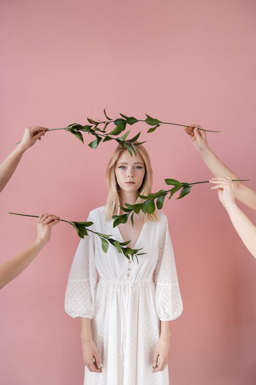 Free A serene portrait of a woman in a white dress surrounded by green leaves against a pink backdrop. Stock Photo