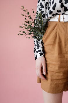 Stylish woman in patterned blouse and shorts holding greenery against a pink backdrop.
