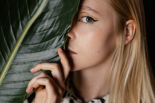 Close-up portrait of a woman with blonde hair and blue eyes holding a large tropical leaf.