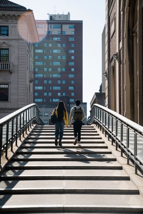 Free A man and woman with backpacks walk up city stairs, sunlight creating a cinematic scene. Stock Photo