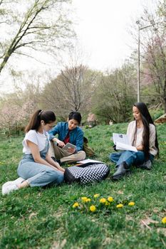 Three young adults studying together on grass in a park surrounded by spring trees.