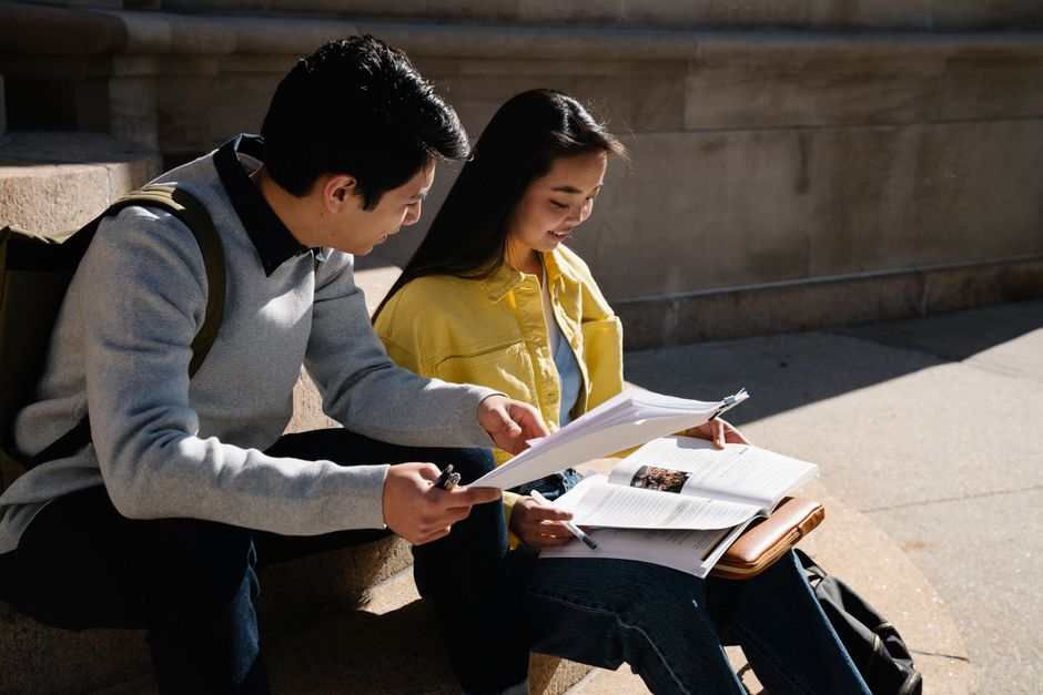 二人の学生が外で一緒に勉強し、書籍やノートを使って読書と議論を行っています。
