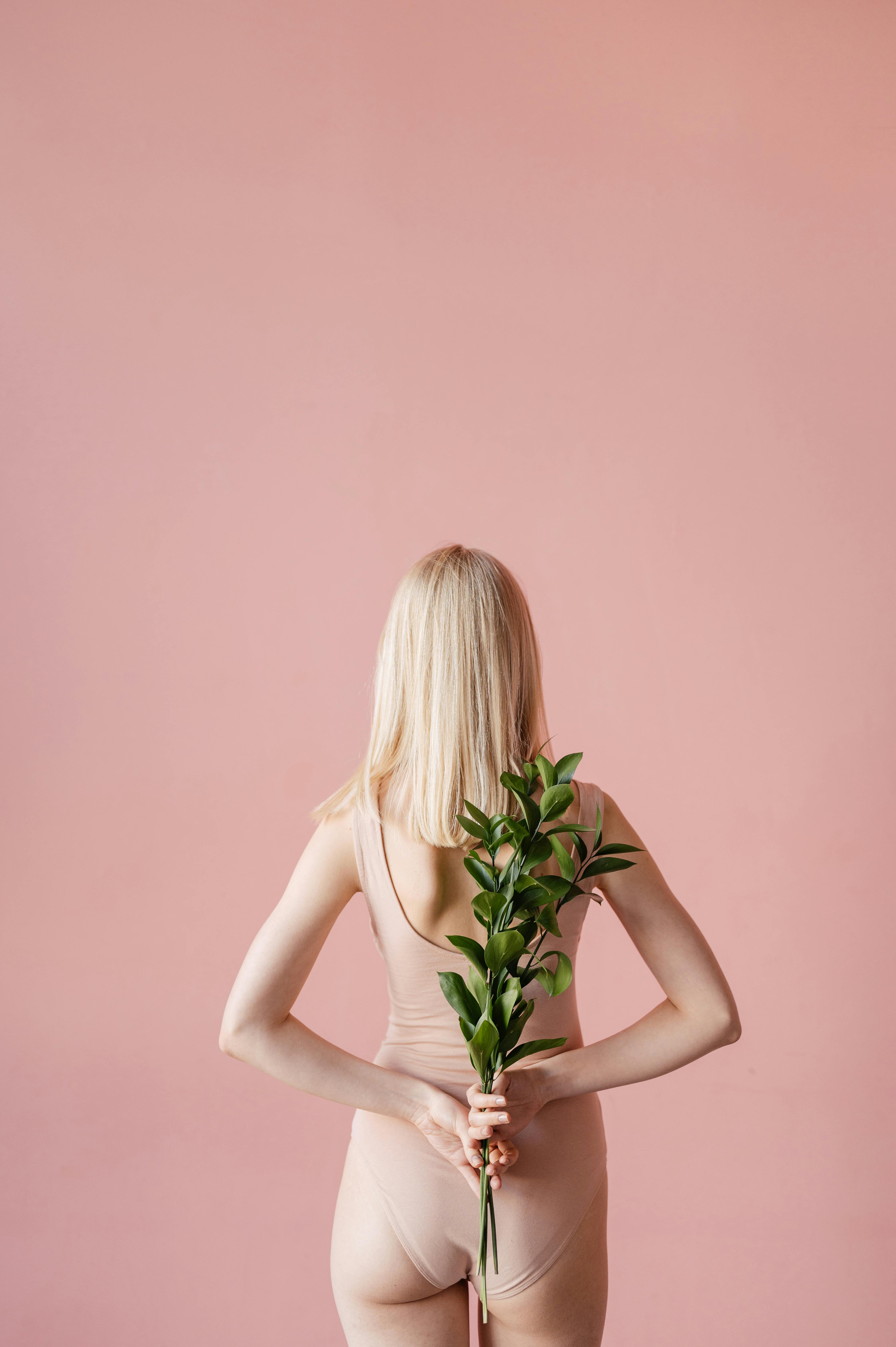 Back view of a woman in nude swimwear holding a green branch against a soft pink studio backdrop.