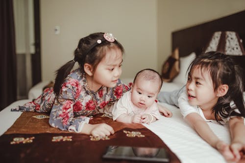 Adorable Asian smiling little girls cuddling newborn baby lying on white delicate blanket on bed in bedroom