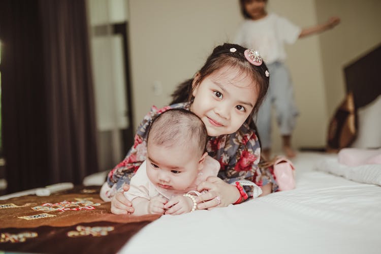 Happy Asian Girl Child Embracing Newborn Baby Lying On Bed