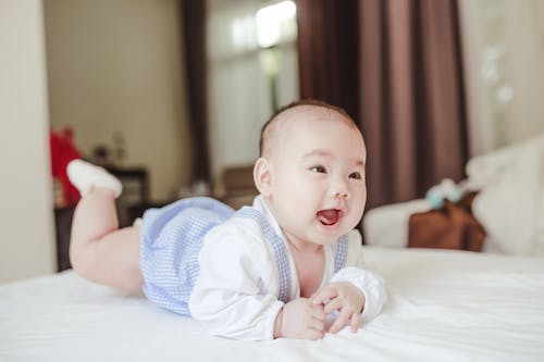 Free Cheerful adorable baby boy in bodysuit and socks lying on belly on white soft blanket on bed Stock Photo