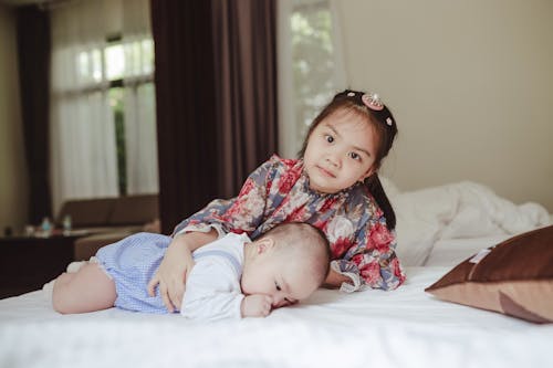 Cute little girl in floral dress embracing newborn baby boy lying on belly on soft blanket on bed in light room