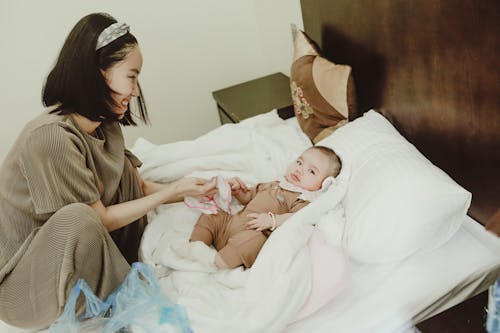 Free Young smiling Asian female mom in home clothes coddling with baby in bodysuit lying on white coverlet on bed Stock Photo