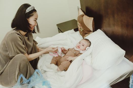 Young smiling Asian female mom in home clothes coddling with baby in bodysuit lying on white coverlet on bed