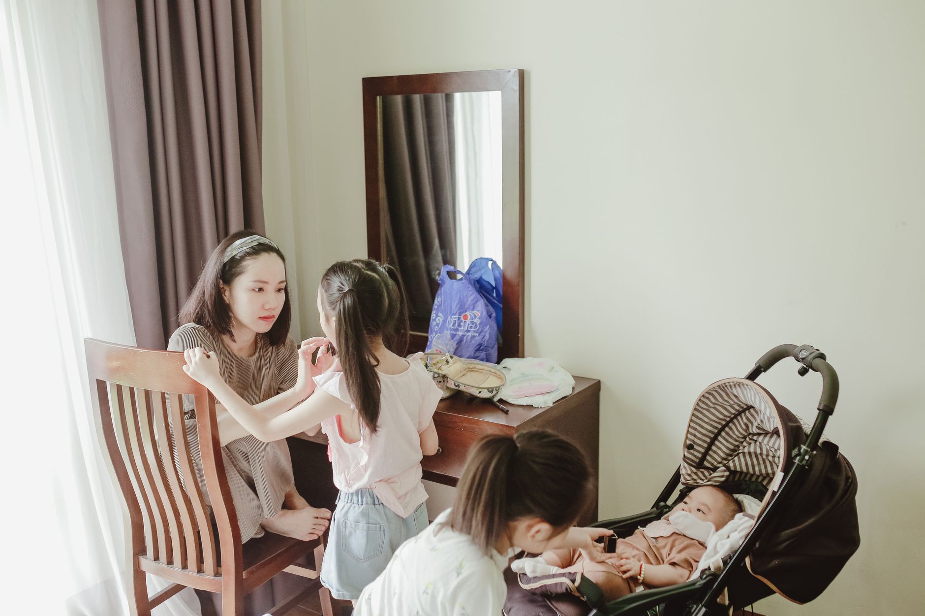 Young delighted Asian female mother coddling preschool daughters and newborn lying in baby stroller in light room
