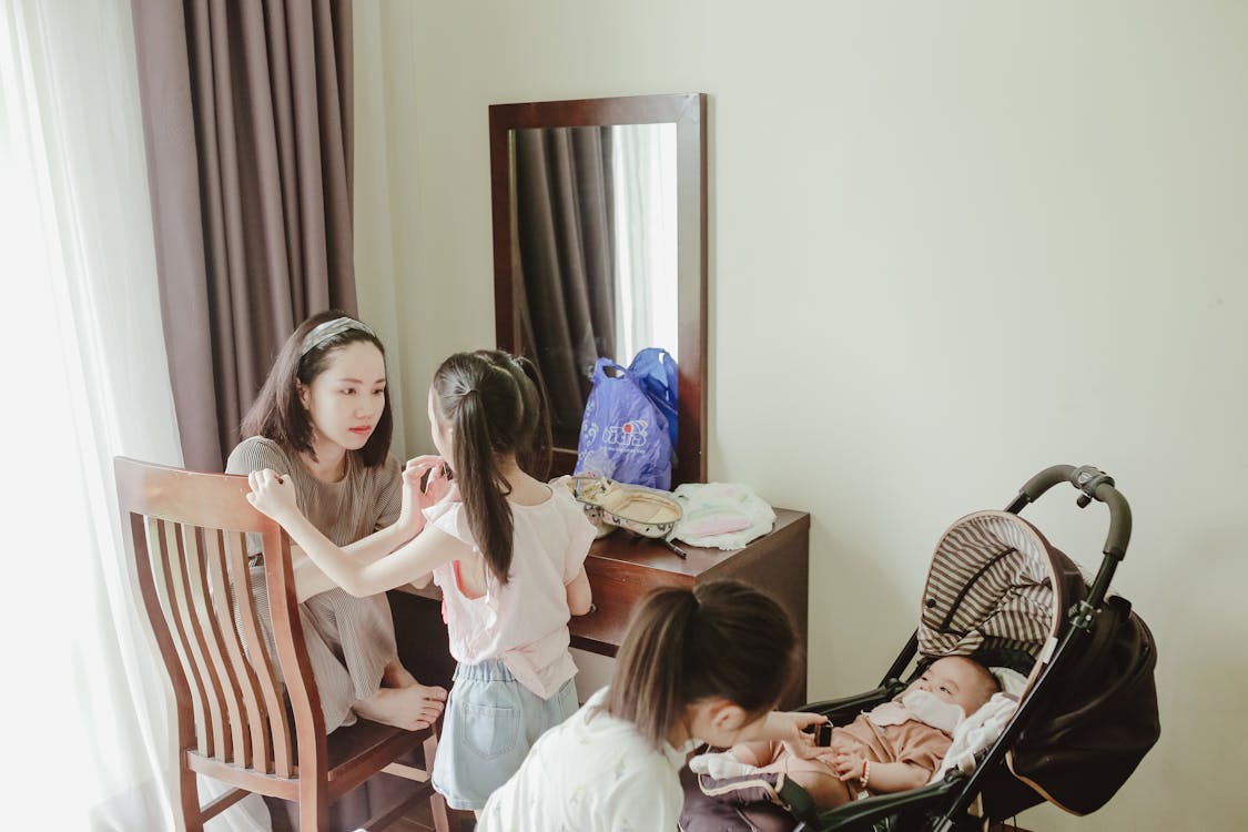Young delighted Asian female mother coddling preschool daughters and newborn lying in baby stroller in light room