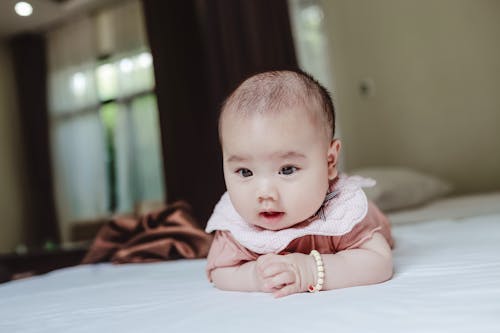 Free Funny baby in casual clothes lying on stomach on bed covered with white sheet in daytime Stock Photo