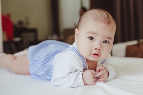 Charming little baby in white and blue pajamas lying on white blanket in bedroom and looking at camera