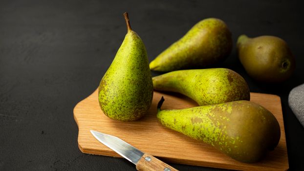 Four ripe green pears on a wooden board with a knife on a dark background.