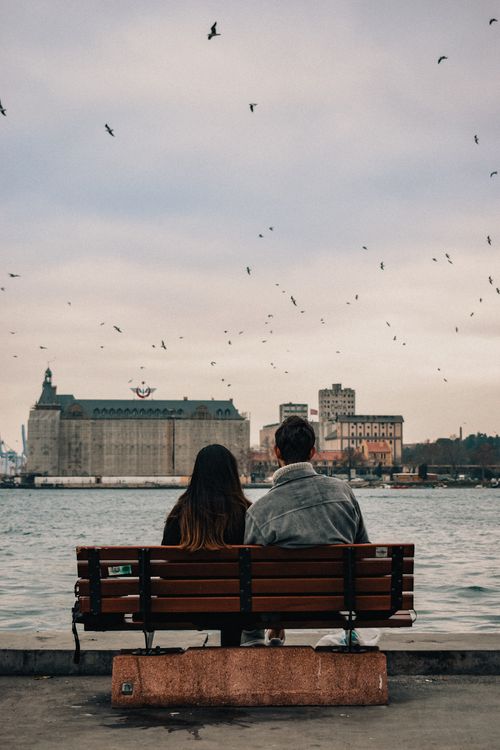 Free A couple enjoys a peaceful moment on a bench by the water, facing an urban landscape. Stock Photo
