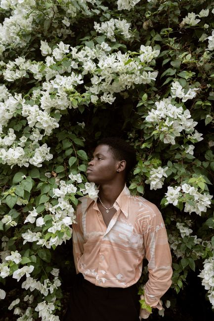 An African American adult man standing by lush white flowers, looking upward contemplatively.