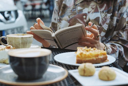 Free Crop unrecognizable female with burning cigarette reading textbook at cafeteria table with pie pieces and cups of coffee Stock Photo
