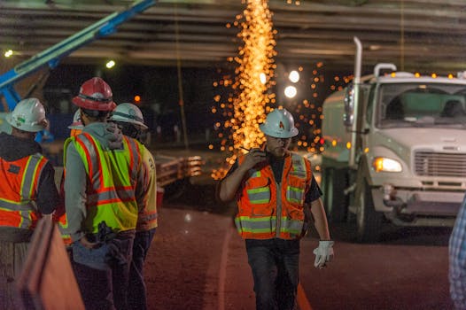 Construction workers in safety gear working at night with sparks flying under a bridge.