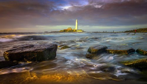 Free A scenic view of a lighthouse surrounded by crashing waves and dramatic skies at sunset. Stock Photo